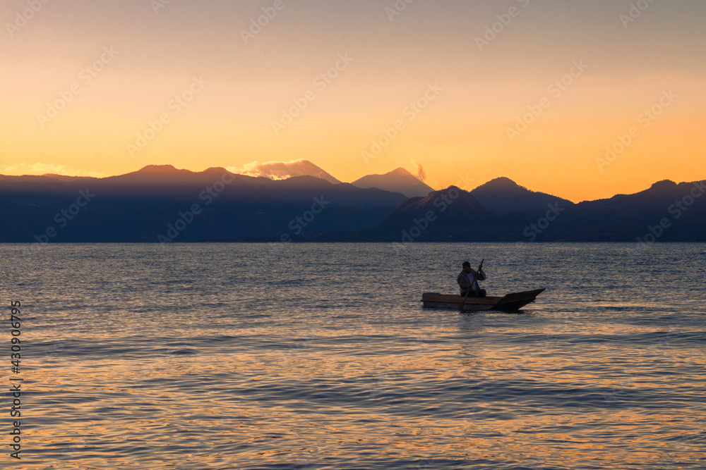 man sailing in a wooden boat on a lake in the middle of a beautiful sunrise with several mountains in the background