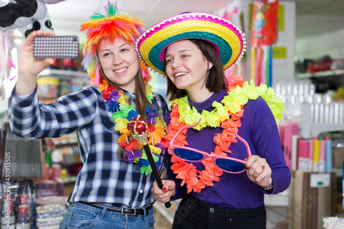 Two cheerful young female friends taking selfie in festive things store while preparing for party