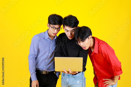 3 adults asian handsome men standing and 1 of them hold laptop on hand. Them look at laptop while feel excited with something on laptop. Shooting in studio with isolated yellow background © Bangkok Click Studio