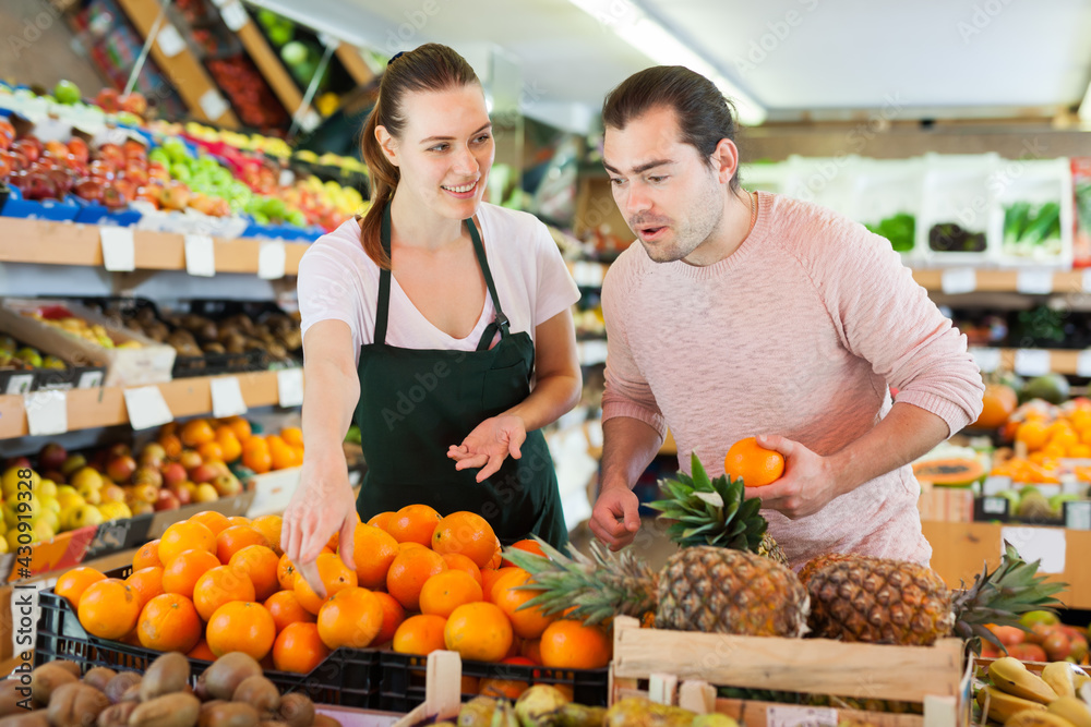 Fine woman wearing apron selling fresh oranges to man customer in fruit store