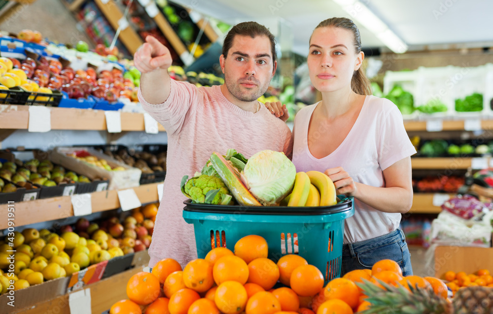 Positive family couple standing with full cart after shopping and pointing to shelves in fruit store