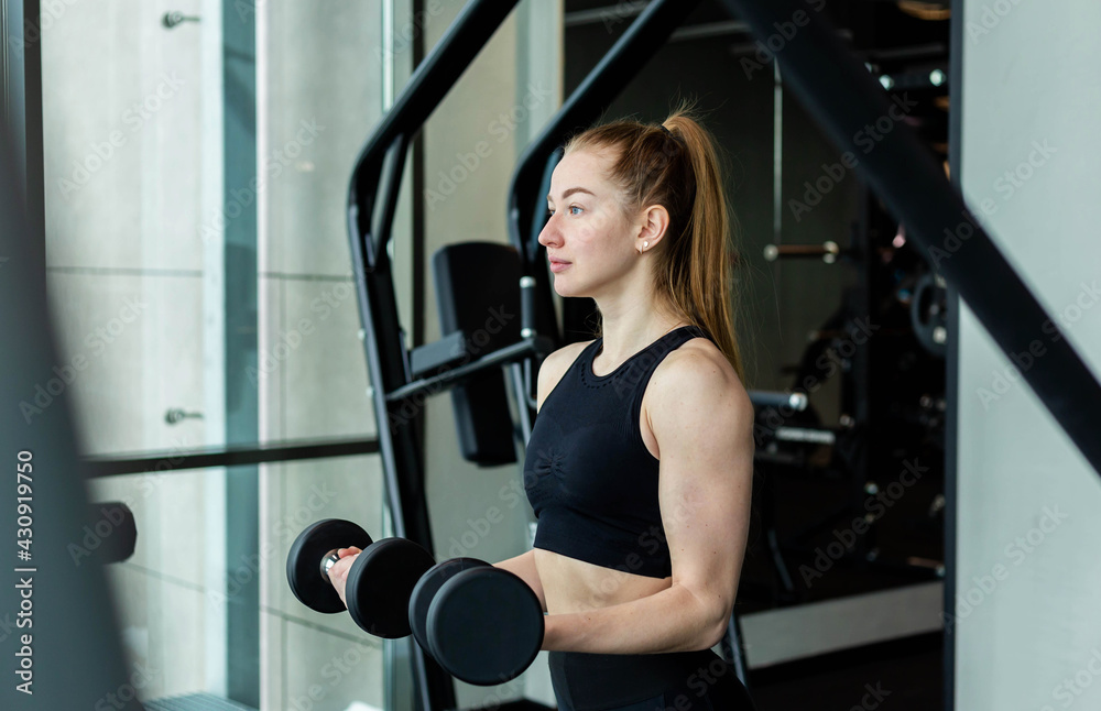 A young girl in black sportswear is engaged in the gym with dumbbells.