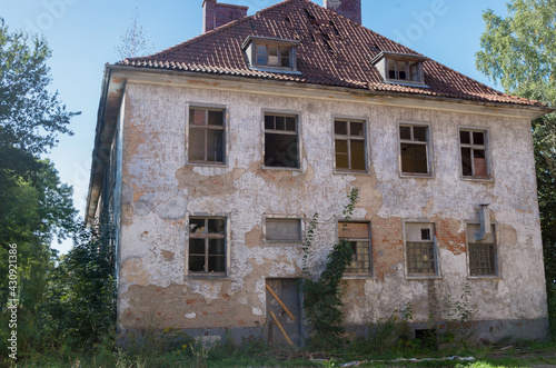 Abandoned dilapidated old brick two-story apartment building with scuffed plaster, broken glass from wooden frames, partially collapsed roof tiles and boarded-up door, with ground overgrown with grass