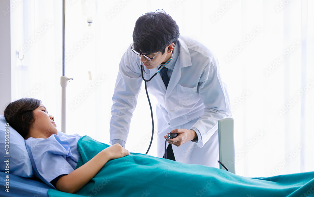 Asian young male doctor wearing eyeglasses and white gown, using stethoscope, checking up health of sick female patient lying down on bed with blanket in ward recovery room in hospital.