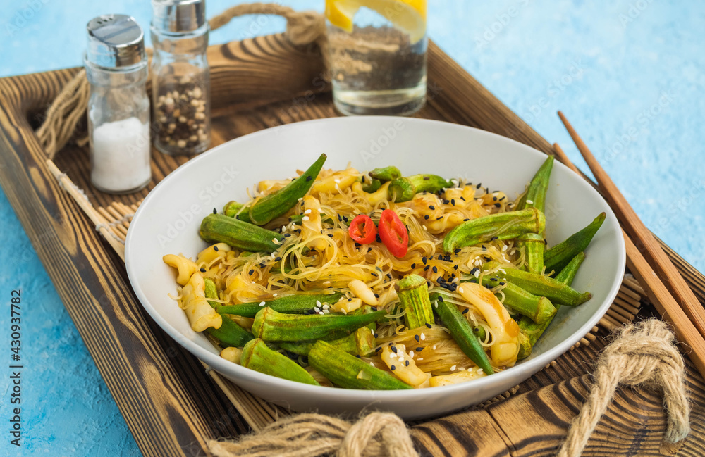 Stir fry, thin rice noodles with squid and okra in a gray plate on a light blue concrete background. Okra recipes.