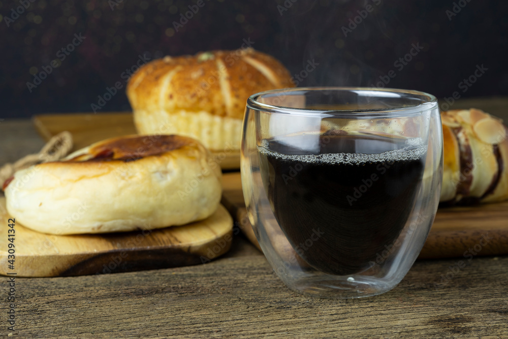 Hot black coffee is placed on the wooden table, there is bread in the background. Hot black coffee is placed on the wooden table, there is bread in the background.