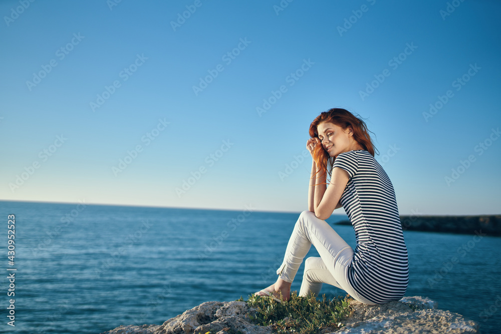 woman in a striped T-shirt and in white trousers in the summer on a stone near the sea in the mountains woman in a striped T-shirt and in white trousers in the summer on a stone near the sea in the mountains