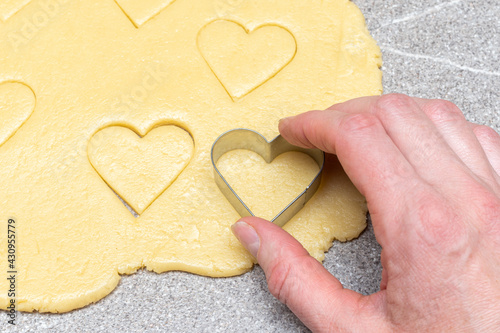 Pastry chef's hand cuts heart-shaped cookies from raw yellow dough on a gray table background, close-up.