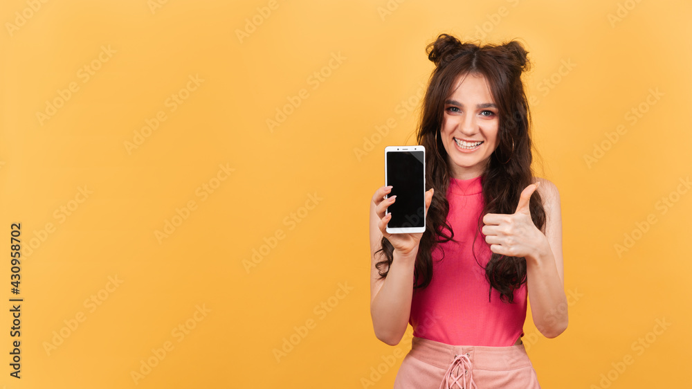 A smiling woman points to the blank screen of her smartphone, standing on a yellow background. A copy of the space. Space for text
