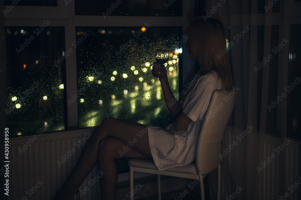 Young slender blonde woman in a white shirt with a glass of red wine sits on a white chair in front of the floor-to-ceiling window and looks out at the night city
