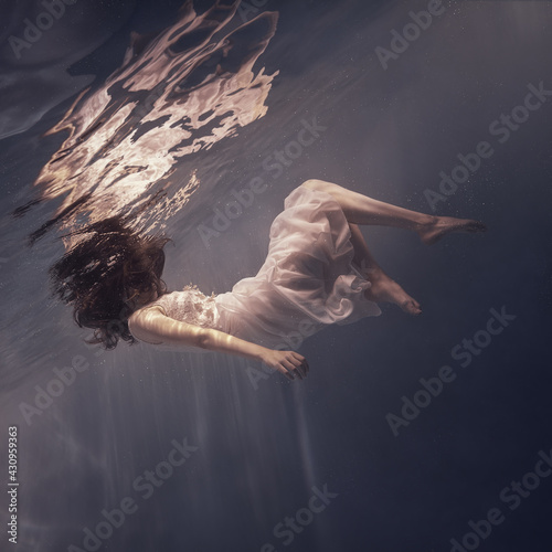 A girl in a white dress with long dark hair swims underwater as if floating in weightlessness