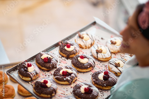 Close up of a tray full of handmade delicious donuts ready for a pastry shop hold by a happy small busines female owner. Pastry, dessert, sweet, making