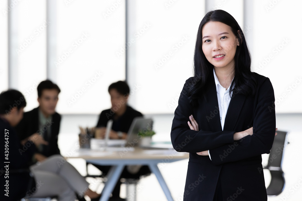 Young Asian businesswoman in black suit standing and posing to camera in office with her team in blur background Young Asian businesswoman in black suit standing and posing to camera in office with her team in blur background