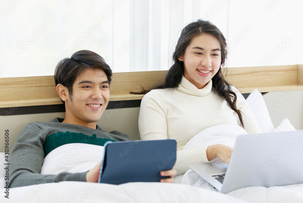 Portrait of cute smiling young Asian lover couple in white and gray long sleeve sweatshirt lying on a bed while using a dark blue digital tablet together. Woman see funny things and laugh out Portrait of cute smiling young Asian lover couple in white and gray long sleeve sweatshirt lying on a bed while using a dark blue digital tablet together. Woman see funny things and laugh out