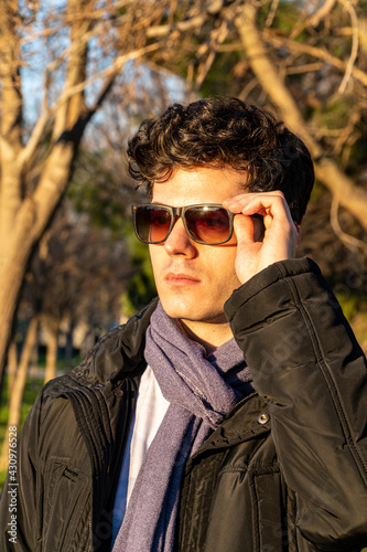 Caucasian curly-haired boy in coat and scarf, putting on sunglasses in a sunset © Javier Paredes