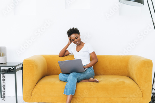 Beautiful young African American woman in white t-shirt and blue jeans using laptop sitting on yellow sofa in bright modern interior