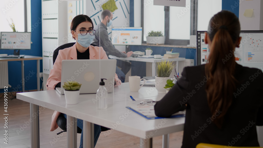 Naklejka premium Freelancer talking with coworker about business startegy while sitting in new normal office wearing protective face mask to prevent infection with coronavirus. Team respecting social distancing