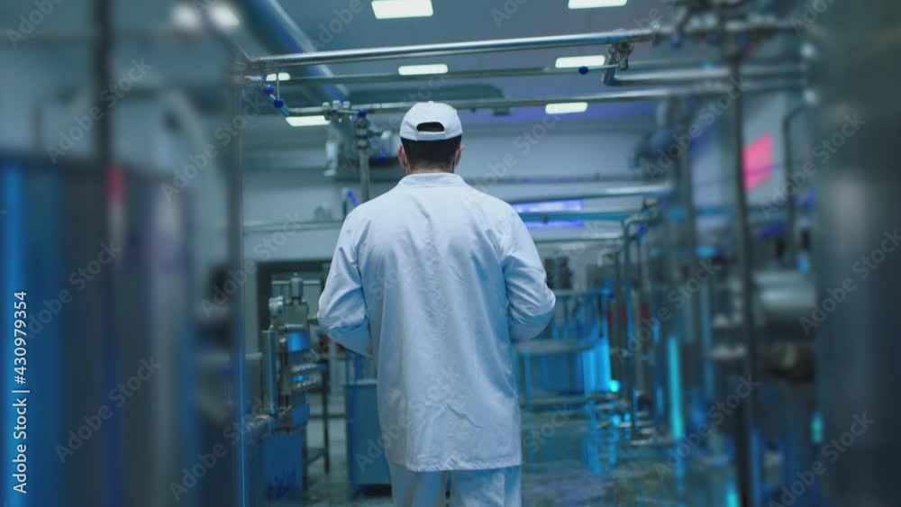 Back rear view of male worker dressed on white uniform standing or walking on modern industrial factory . Man using tablet and doing notes inside milk or juice factory with metal tanks . Slow motion Back rear view of male worker dressed on white uniform standing or walking on modern industrial factory . Man using tablet and doing notes inside milk or juice factory with metal tanks . Slow motion