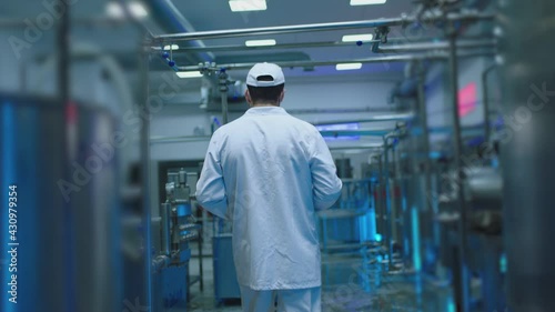 Back rear view of male worker dressed on white uniform standing or walking on modern industrial factory . Man using tablet and doing notes inside milk or juice factory with metal tanks . Slow motion
