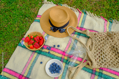 still life ripe strawberries in straw plate, straw hat, sunglasses, empty glasses, and food on checkered plaid on grass. top view. summer picnic in the park. Copy space
