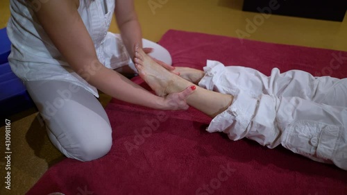 Woman wearing white clothes massaging feet of female lying on mat. Partial view Thai spa therapist doing relaxational and healing massage. Concept of wellness photo