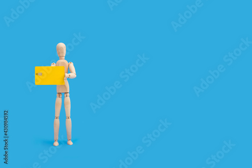 Muñeco maniqui de madera sosteniendo una tarjeta de crédito amarilla sobre un fondo azul liso y aislado. Vista de frente. Copy space. 