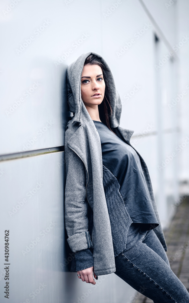 Beautiful girl in a hood with long dark hair in a gray sweatshirt with a black leather backpack near a gray wall in an industrial zone of a factory Beautiful girl in a hood with long dark hair in a gray sweatshirt with a black leather backpack near a gray wall in an industrial zone of a factory