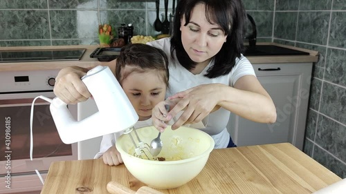 Mother with smile teaching little daughter cooking dough: the girl puts cocoa into the dough and licks a spoon. Preparation for the holiday, help in the kitchen