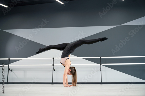 Side view of strong fit woman wearing sportswear standing on arms and practicing split in air. Flexible female dancer training before competition in dance hall with ballet handrails, hi tech interior.