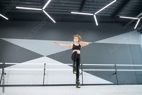 From below view of active female teenager doing dance cardio workout in hall with hi tech interior. Young pretty girl wearing black sportswear practicing dance moves while doing kangoo jumps.