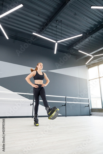 Young caucasian girl with ponytail practicing kangoo jumping indoors. From below view of female athlete holding hands on waist  enjoying cardio dance workout in hall  hi tech interior.