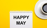 On a bright yellow background, a white cup with coffee and a white notepad with the words HAPPY MAY