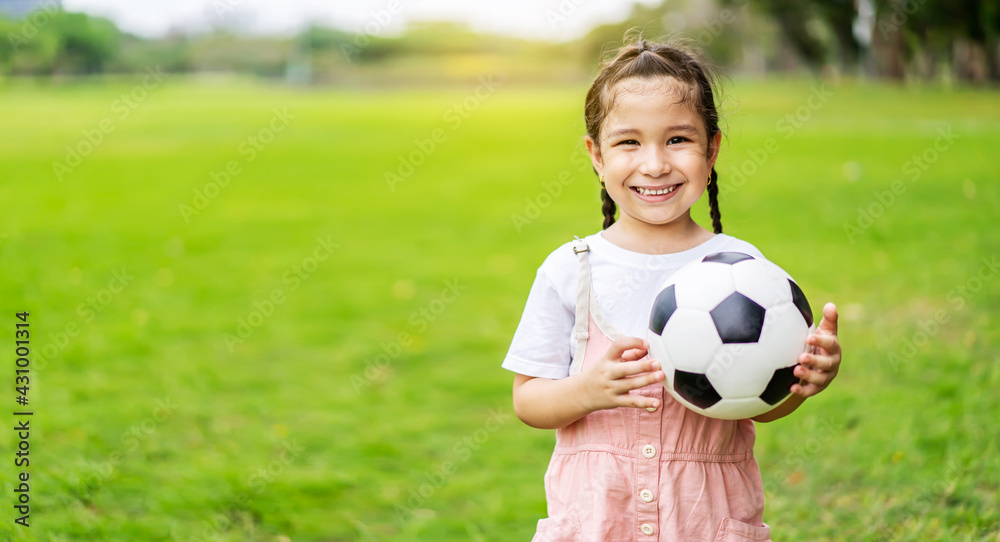 Smiling little girl holding soccer ball standing at green football field in summer day. Portrait of little girl athlete playing with a ball at stadium. Active childhood concept. Copy space