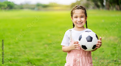 Smiling little girl holding soccer ball standing at green football field in summer day. Portrait of little girl athlete playing with a ball at stadium. Active childhood concept. Copy space © TeTe Song