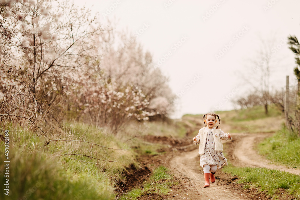 Little girl in rubber boots with two braids, running on a path in countryside on spring day.