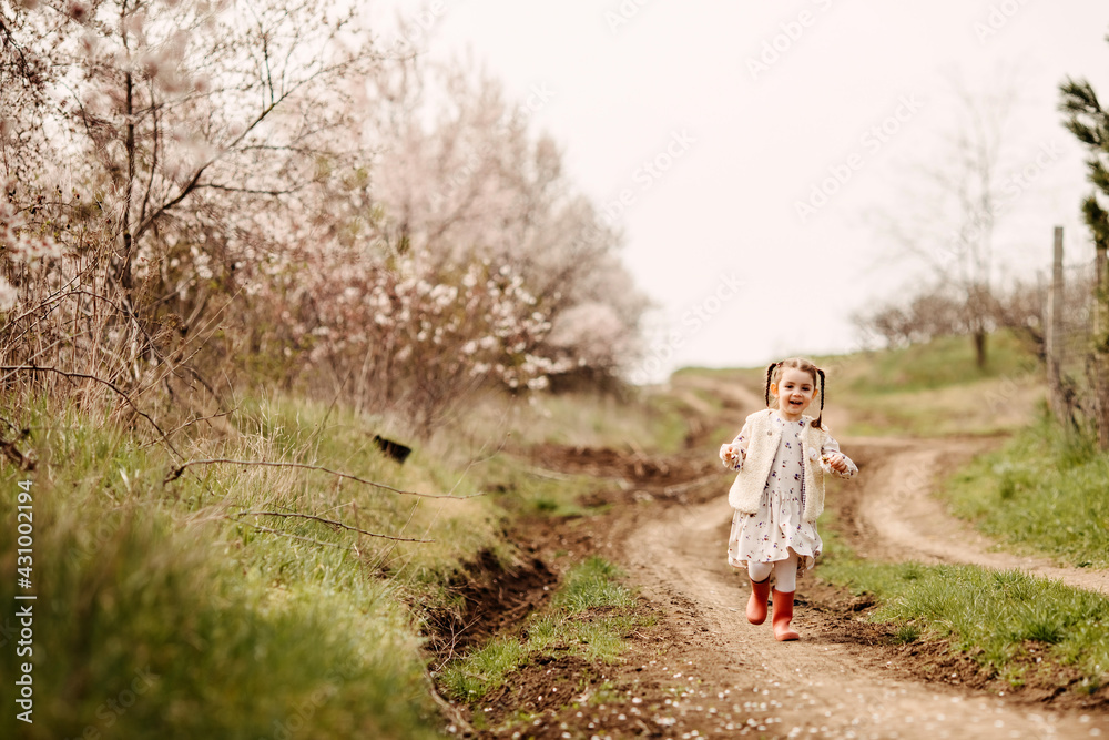 Happy little girl in rubber boots with two braids, running on a path in countryside on spring day.