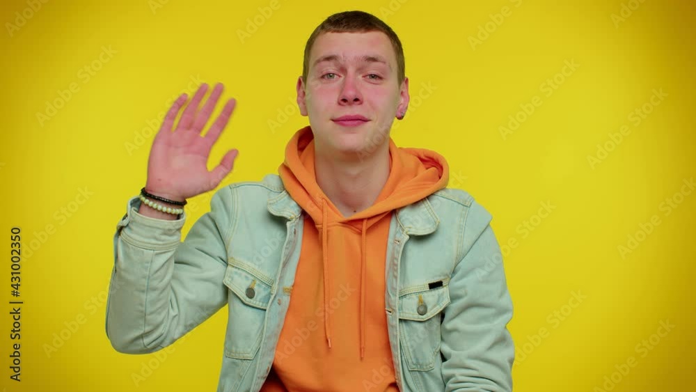Handsome teenager boy smiling friendly at camera and waving hands gesturing hello or goodbye, welcoming with hospitable expression. Young fashionable man indoors isolated on yellow wall background Handsome teenager boy smiling friendly at camera and waving hands gesturing hello or goodbye, welcoming with hospitable expression. Young fashionable man indoors isolated on yellow wall background