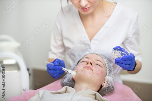 Cosmetology and acne treatment. Professional woman dermatologist in rubber gloves, applies the mask to the client's face with cosmetology brush. Young teen girl with problem skin lying on the couch