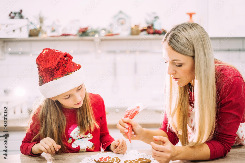 Mom and daughter in the kitchen glaze on gingerbread cookies in red sweaters and Santa hats with lights in the background. Christmas preparations