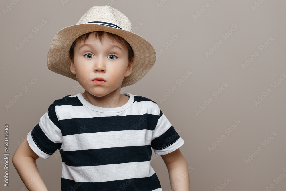 attractive toddler in a straw hat and striped T-shirt looks at the camera in surprise, a studio portrait of a child on an isolated bearish background. attractive toddler in a straw hat and striped T-shirt looks at the camera in surprise, a studio portrait of a child on an isolated bearish background.