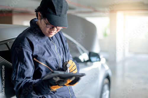 Asian auto mechanic holding digital tablet checking car in garage. Mechanical maintenance engineer working in automotive industry. Automobile servicing and repair concept