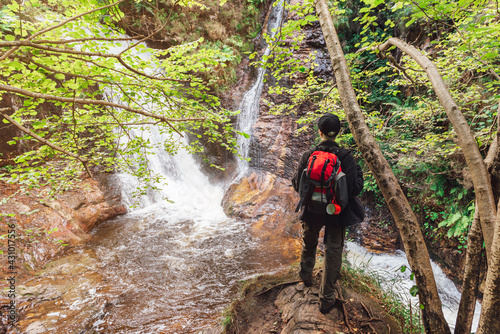 hiker on back-turned hiker with red backpack standing on a rock contemplating a waterfall in a jungle setting. Person enjoying the forest on a weekend getaway. active tourism and adventure activities.
