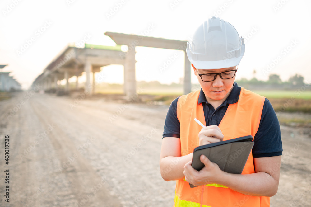 Smart Asian worker man or male civil engineer with protective safety helmet and reflective vest using digital tablet for project planning and checking architectural drawing at construction site. Smart Asian worker man or male civil engineer with protective safety helmet and reflective vest using digital tablet for project planning and checking architectural drawing at construction site.