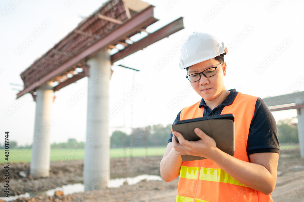 Fototapeta premium Smart Asian worker man or male civil engineer with protective safety helmet and reflective vest using digital tablet for project planning and checking architectural drawing at construction site.