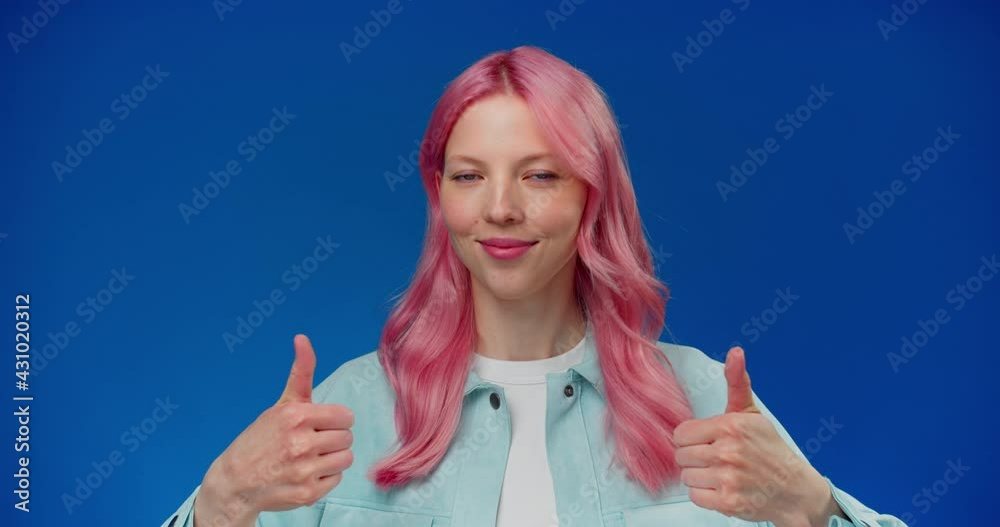 Beautiful 20s female with pink hair raising thumb up, smiling broadly, laughing at camera, showing cool, like, success gesture on blue background.Young woman nods approvingly and makes a cool gesture Beautiful 20s female with pink hair raising thumb up, smiling broadly, laughing at camera, showing cool, like, success gesture on blue background.Young woman nods approvingly and makes a cool gesture