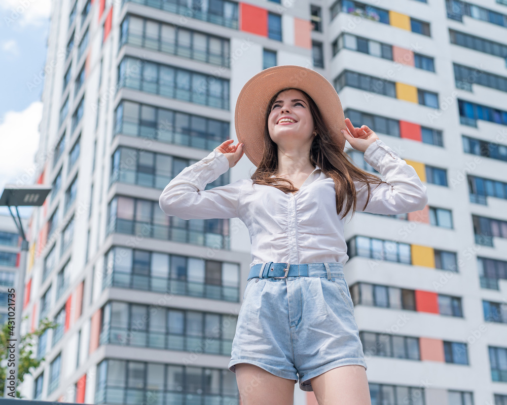 Happy caucasian young woman in a hat and shorts walks on a city street on a hot summer day - obrazy, fototapety, plakaty 