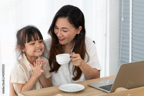 Young little girl bring cup of coffee to her mother, while young mother working on desk using laptop computer during work from home. Mom working at home with her child bring her cup of coffee.