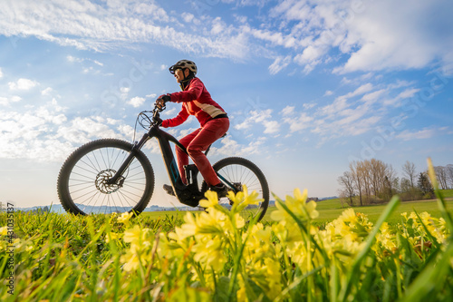 pretty mid age woman riding her electric mountain bike in early springtime in the Allgau mountains near Oberstaufen, in warm evening light with blooming spring flowers in the Foreground