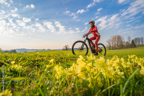 pretty mid age woman riding her electric mountain bike in early springtime in the Allgau mountains near Oberstaufen, in warm evening light with blooming spring flowers in the Foreground