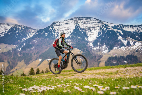 pretty mid age woman riding her electric mountain bike in early springtime in the Allgau mountains near Oberstaufen,below the spectacular snow capped mount HochgratNagelfluh chain